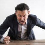 Human Error - Expressive angry businessman in formal suit looking at camera and screaming with madness while hitting desk with fist